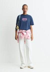 Junge Frau mit geflochtenem Haar, die ein navyblaues "Wrong Button"-T-Shirt, weiße Hosen, ein um die Taille gebundenes pinkes Hemd trägt und eine avocadoförmige Tasche trägt.