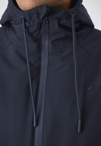 Marineblaue Kapuzenjacke aus glattem, leichtem Material mit einem Frontreißverschluss, elastischen Zugbändern und einem dezenten Logo an der Seite.