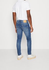 Jeans de denim azul ajustados con cintura de altura media, bolsillos traseros y un parche de cuero marrón en la cinturilla, combinados con zapatillas blancas.