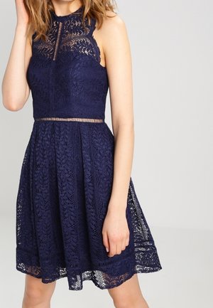 Mouwloze marineblauwe kanten jurk met hoge halslijn, nauwsluitend lijfje en uitlopende rok, gedragen door een staand persoon tegen een effen achtergrond.