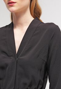 Svart omlottklänning i mjukt tyg, med V-ringning, långa ärmar och subtila sömdetaljer längs kanterna.