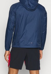 Chaqueta cortavientos marina con capucha, que presenta una textura suave y puños elásticos. Los detalles incluyen un logo cerca del dobladillo y un diseño sencillo.