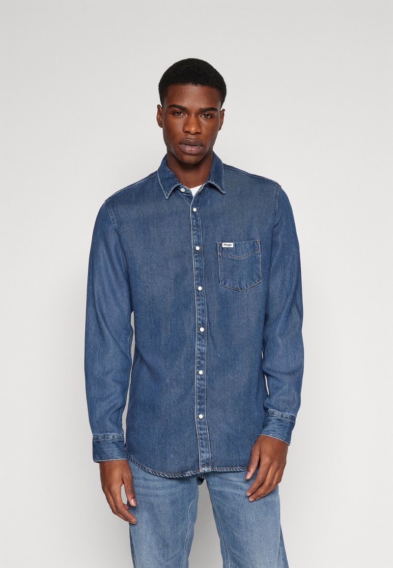 Chemise en denim à manches longues de couleur bleu foncé, avec une fermeture à boutons à l'avant, une poche poitrine unique et des coutures contrastées. Tissu doux et texturé.