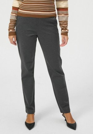 Žena u tamnosivim hlačama ravnih nogavica, smeđem prugastom džemperu i crnim cipelama s špicom stoji na bijeloj pozadini.