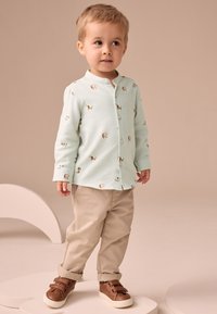 Camisa de manga larga verde claro con estampado de perro, abotonada al frente; pantalones beige; zapatillas marrones con velcro; texturas sencillas y lisas.