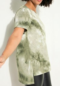 Camiseta tie-dye en verde oliva con mangas cortas, que presenta un corte relajado y aberturas laterales para mayor movilidad. Textura de algodón suave.