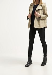 Beige trenchcoat med knappdetaljer, bärs över en marinblå topp, kombineras med svarta smala jeans och ankelskor, stående mot en neutral bakgrund.