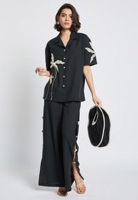 Camicia di lino nera con maniche corte e bottoni frontali, caratterizzata da ricami floreali. Abbinata a pantaloni coordinati con fessure laterali.