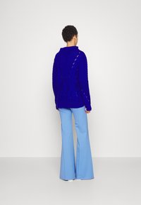 Personne debout, portant un pull tricoté épais bleu royal avec des motifs torsadés et un pantalon large bleu clair sur un fond uni.