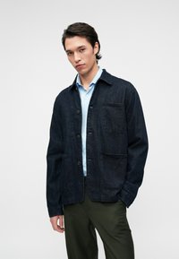 Chaqueta de mezclilla oscura con frente abotonado, dos bolsillos frontales y cuello con solapa, combinada con una camisa azul clara y pantalones verde oliva.