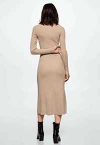 Robe longue beige côtelée avec manches longues et silhouette ajustée, se terminant au milieu du mollet. Associée à des bottines noires à talons moyens.