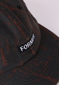 Șapcă neagră cu un model marmorat maro, cu etichetă brodată albă "FORMER" pe partea din față. Material din bumbac cu suprafață texturată.