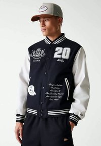 Jovem vestido com uma jaqueta varsity em preto e branco com os logótipos "20" e New Era, combinada com um boné bege da New Era, olhando para o lado.
