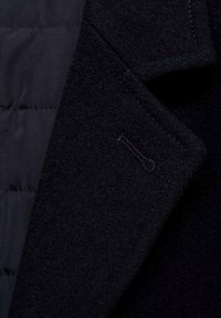 Primo piano di un cappotto blu scuro con un esterno in lana texturizzata, colletto sagomato e colore uniforme con dettagli di cucitura visibili sulla revers.