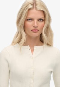 Prążkowany, kremowy top z długim rękawem, z okrągłym dekoltem i pięcioma guziczkami z przodu. Tkanina wydaje się miękka i fakturowana.