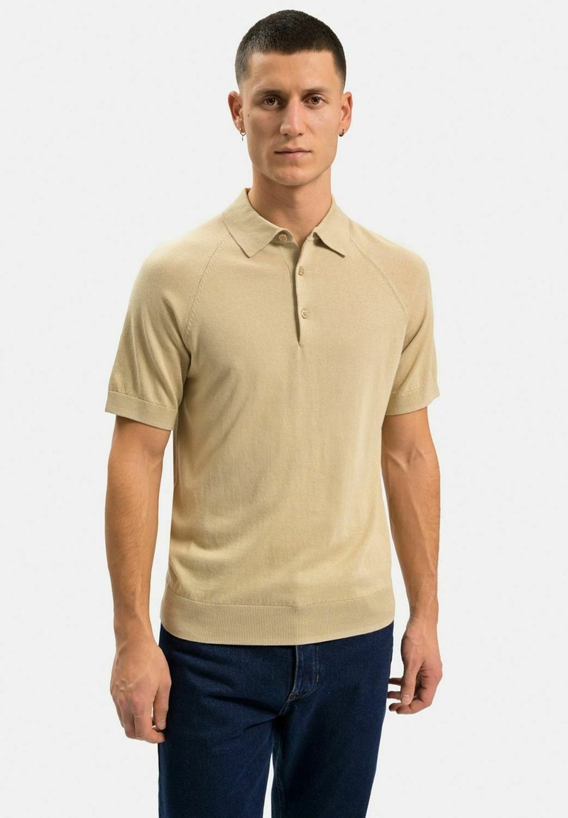 Beige kortärmad polotröja med mjuk textur, treknapplåda och ribbade fållar som bärs över mörkblå jeans.