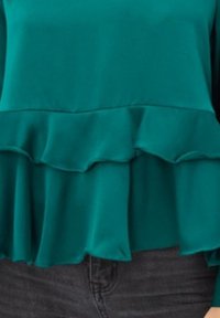 Blusa verde azulado con un diseño de volantes en capas en el dobladillo, hecha de una tela suave. Combinada con jeans ajustados gris oscuro.