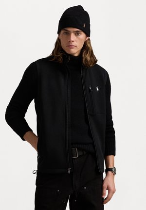 Sort vest med høj krave, lynlåslukning og lomme på venstre bryst. Båret over en sort turtleneck, parret med mørke bukser og en strikket beanie.