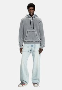 Hoodie gris avec une poche avant et des cordons, associé à un jean large bleu clair et des baskets grises avec des accents rouges.