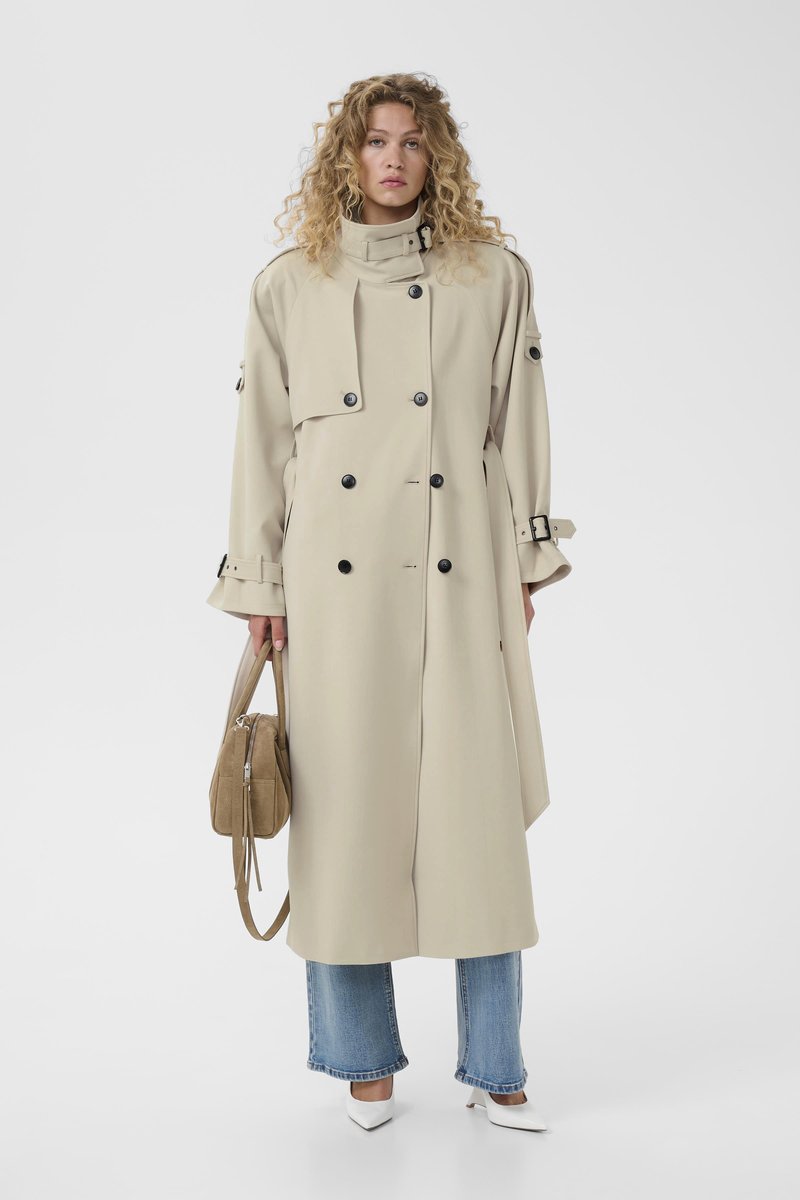 Femme aux cheveux bouclés portant un trench-coat beige à double boutonnage, un jean bleu, des talons blancs, et tenant un sac à main marron clair.