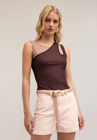 Ruskea ja beige epäsymmetrinen tankkitoppi, jossa on verkko-osio ja olkaimellinen leikkaus, yhdistettynä vaaleanpinkuisiin shortseihin ja yhteensopivaan vyöhön.
