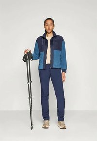 Giacca in pile blu e navy con tasche frontali, abbinata a pantaloni navy. Il modello tiene in mano un bastone da trekking nero, indossando scarpe di colore beige chiaro.