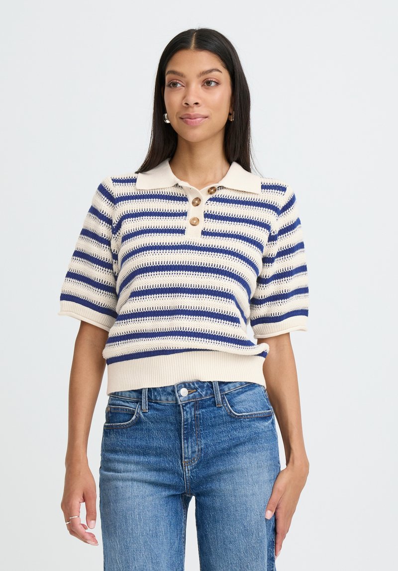 Polo pulover s kratkimi rokavi v mornarsko modri in kremni vodoravni črtasti vzorec, z rebrastim ovratnikom in zapenjanjem na tri gumbe. Nosijo ga s pridihom modrimi kavbojskimi hlačami.