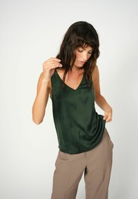 Green satin sleeveless top with a V-neck and a smooth texture, paired with tan trousers. The top drapes softly over the body.