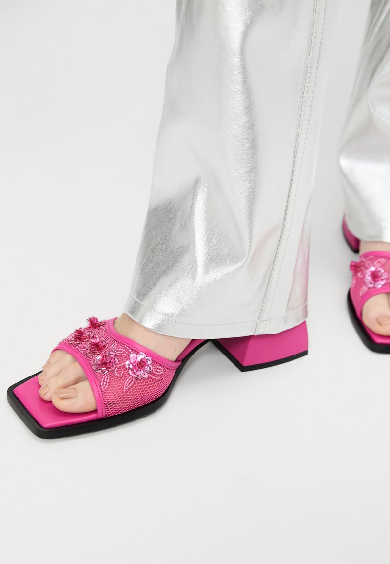 Sandalele cu toc roz au broderie florală și paiete pe un material din plasă, cu vârf pătrat și toc gros. Sunt asociate cu pantaloni argintii strălucitori.