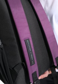 Zaino nero con spallacci viola, con un'etichetta rettangolare con la scritta "PLANET WATER." Tessuto testurizzato e dettagli imbottiti.