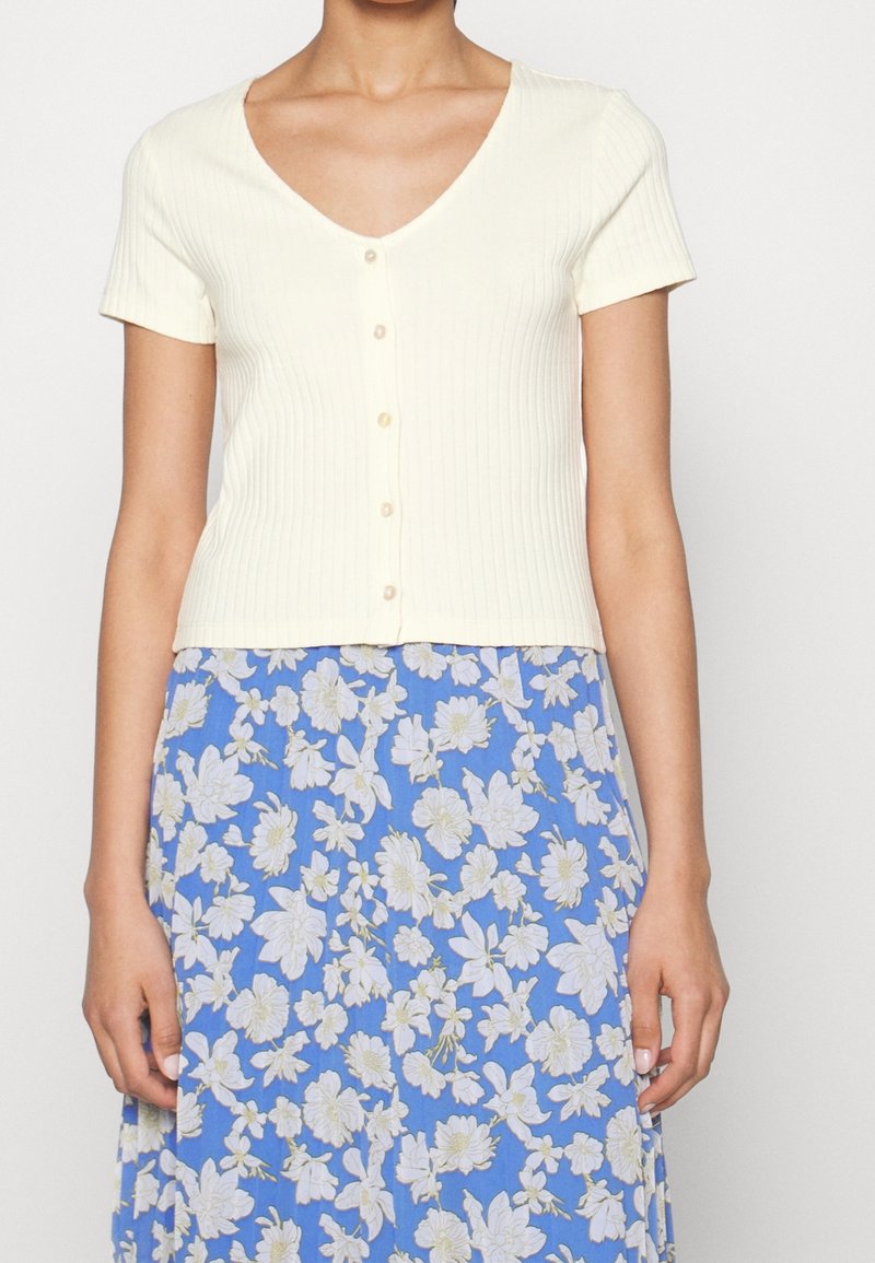 Kvinna iklädd en kortärmad, krämfärgad ribbad topp med knappar, kombinerad med en blå kjol med vit blommönster.
