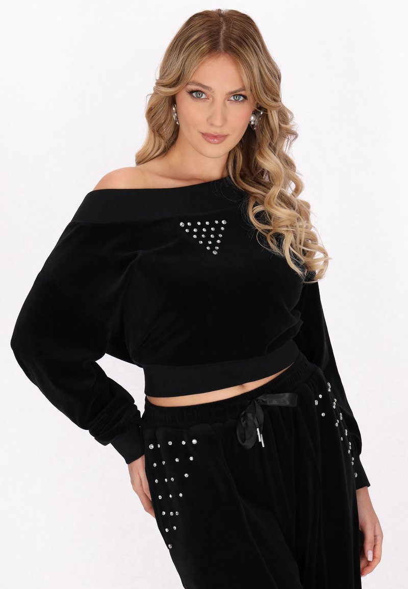 Top off-shoulder in velluto nero con design cropped, caratterizzato da un cluster triangolare di strass. Abbinato a pantaloni neri decorati con strass.