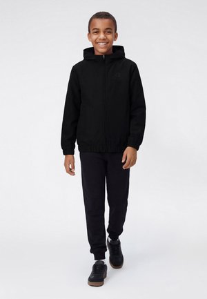 Jongen die lacht terwijl hij loopt, gekleed in een zwarte capuchonjas, zwarte broek, witte sokken en zwarte sneakers tegen een witte achtergrond.