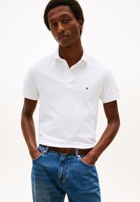 Hvit polotrøye med korte ermer og krage, med en liten brodert logo på brystet. Kombinert med blå denimjeans og et brunt belte.