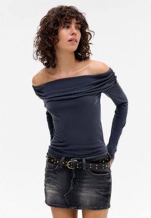 Off-the-shoulder long-sleeve top in dark blue, featuring draped fabric and a fitted silhouette, paired with a black denim skirt and studded belt.