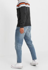 Pull gris avec des rayures orange, blanche et bleue sur la poitrine, associé à un jean bleu clair et des baskets blanches. Coupe décontractée, poignets côtelés.