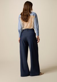 Mujer con cabello largo y castaño, vistiendo una camisa azul con rayas y un panel trasero transparente de encaje, y pantalones oscuros de pierna ancha, de perfil.