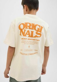 Krémové tričko s oranžovým grafickým textem na zadní straně, na kterém je napsáno "ORIGINALS" a další značkové detaily. Krátké rukávy, volný střih.
