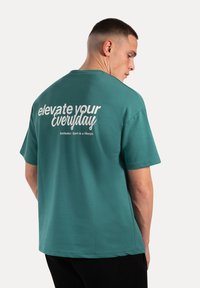 Camiseta de algodón color verde azulado con un corte relajado. Presenta texto blanco "eleva tu día a día" en la parte posterior. Manga corta y escote redondo.