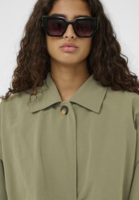 Olive zeleno jakno s ovratnikom in enim gumbom, v kombinaciji z velikimi črnimi sončnimi očali z gradientnimi lečami.
