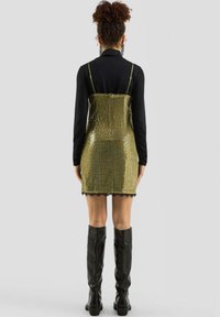 Frau mit lockigem Haar trägt ein schwarzes Langarmoberteil unter einem goldenen Pailletten-Mini-Kleid und schwarzen kniehohen Stiefeln, steht mit dem Rücken zur Kamera.