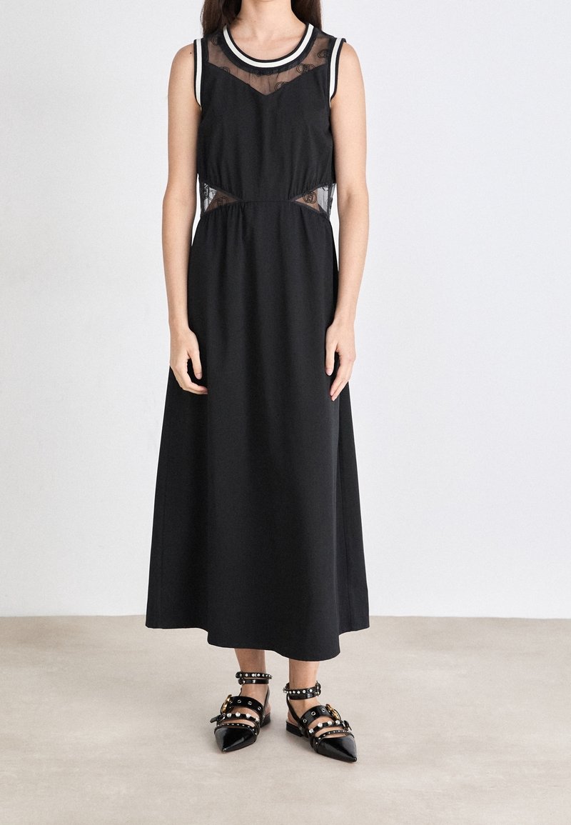 Femme portant une robe midi noire sans manches avec des panneaux en mesh transparent au niveau du cou et de la taille, associée à des ballerines noires à bouts pointus cloutées.