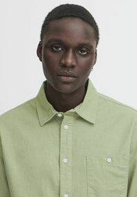Light green button-up shirt with a collar, front pocket, and white buttons. Smooth texture and relaxed fit. Plain design with no patterns.