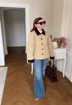 Frau in beiger Jacke mit dunklem Kragen und Bündchen, blauen Jeans, Sonnenbrille, trägt eine dunkle Handtasche und steht in einem Raum mit Holzboden und Blumen.