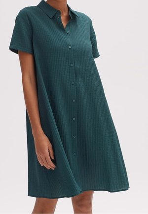 Kobieta ubrana w zieloną, teksturowaną sukienkę zapinaną na guziki z kołnierzykiem i krótkim rękawem, stojąca na tle jednolitego jasnego tła.