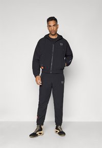 Sort vindjakke med hætte, lynlåslukning, elastisk talje og logodetalje. Sammen med matchende sorte bukser og sporty sorte og brune sko.