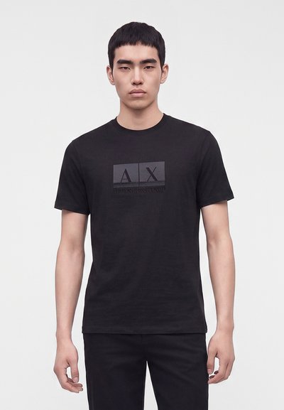 Млад мъж стои лице напред, облечен в черна къс ръкав тениска Armani Exchange с лого на гърдите на едноцветен фон.