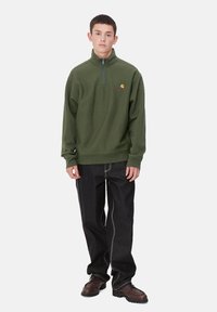 Pulover de forro polar verde oliva con cuello de media cremallera, que presenta un pequeño logo en el pecho. Jeans de mezclilla oscura con costuras blancas en contraste.