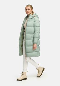 Minkštintas parka paltas šviesiai žalios spalvos, su kapišonu, užtrauktuku, šoninėmis kišenėmis ir elastingais rankogaliais. Suderintas su kreminėmis kelnėmis ir smėlio spalvos batais.