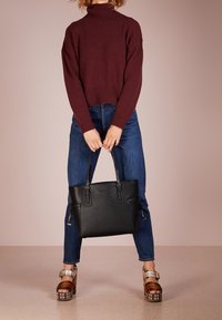 Zwarte leren tote bag met twee bovenhandvatten, rechthoekige vorm en ritssluiting. Ggedragen met een bordeauxkleurige coltrui en blauwe spijkerbroeken.